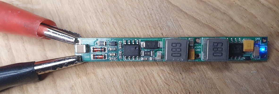 The narrow DC/DC power supply board, showing the input terminals under test clips, two shielded inductors, and the regulated 12 V, 5 V, and ground output pads