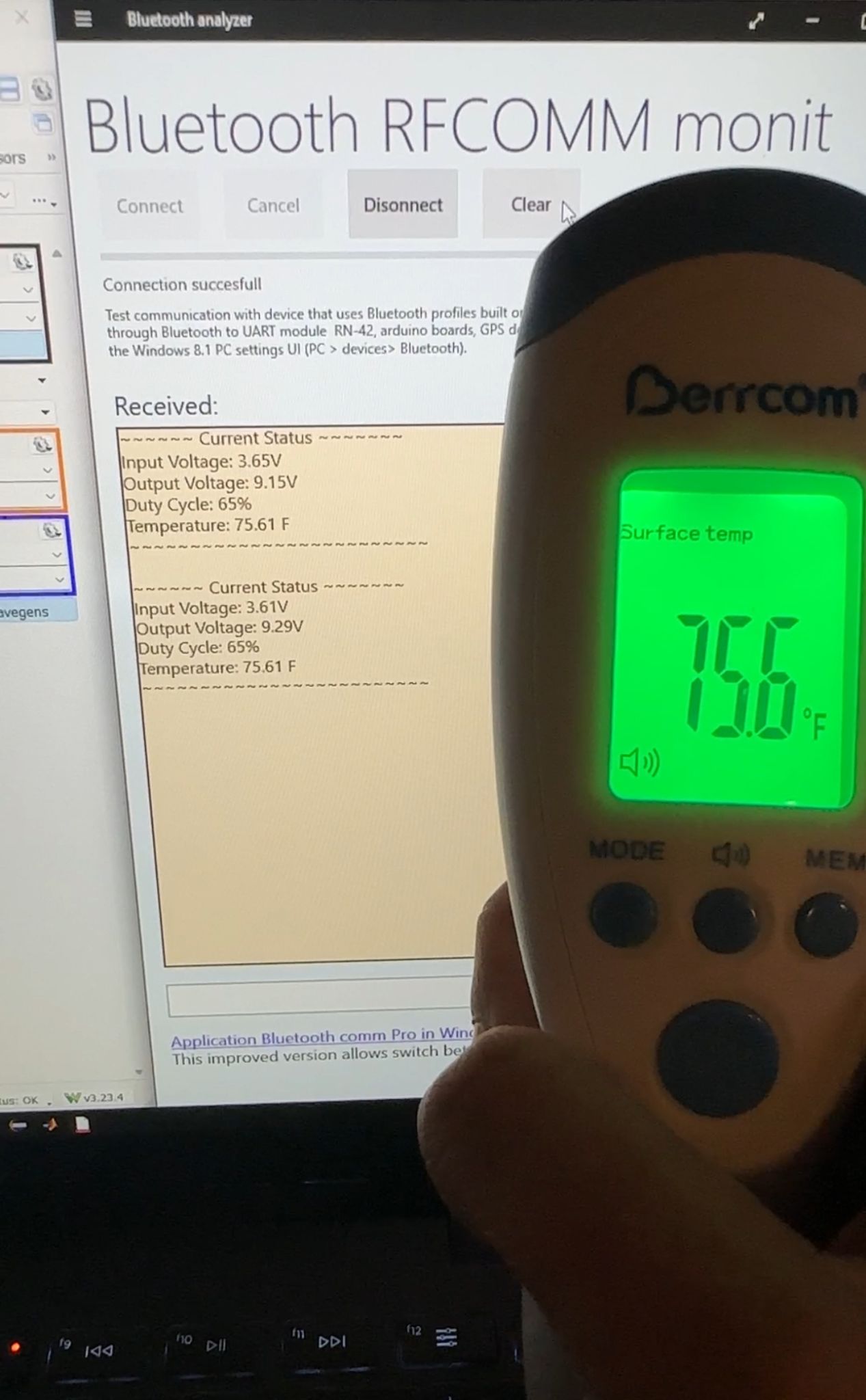A laptop showing received Bluetooth status lines with input voltage, output voltage, duty cycle, and a temperature of 75.61 °F, next to a commercial infrared thermometer reading 75.6 °F